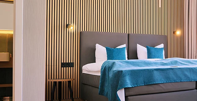 Modernes Hotelzimmer im H24 Hotels mit türkisfarbenen Akzenten und Holzlamellenwand hinter dem Bett.