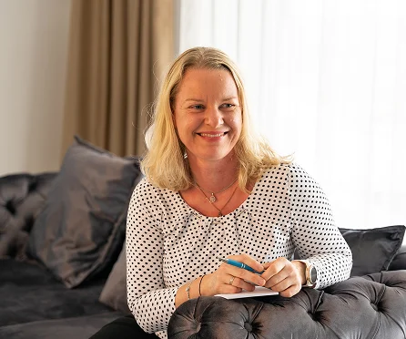 Jeannette sitzt mit Notizblock auf dem Sofa und lächelt freundlich in einem Gespräch