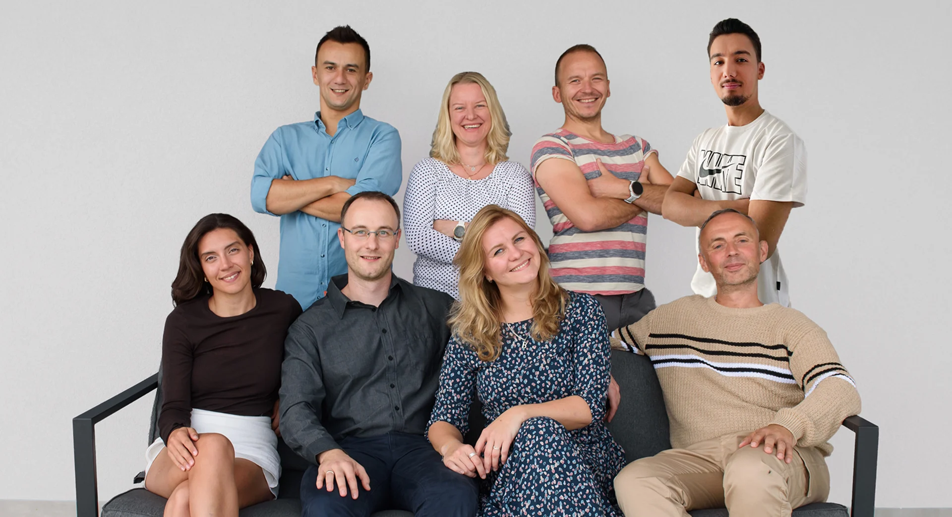 Teamfoto der SEO Revolution GmbH: Sieben lächelnde Teammitglieder, vier stehend im Hintergrund und vier sitzend auf einer grauen Bank vor einer hellen Wand.