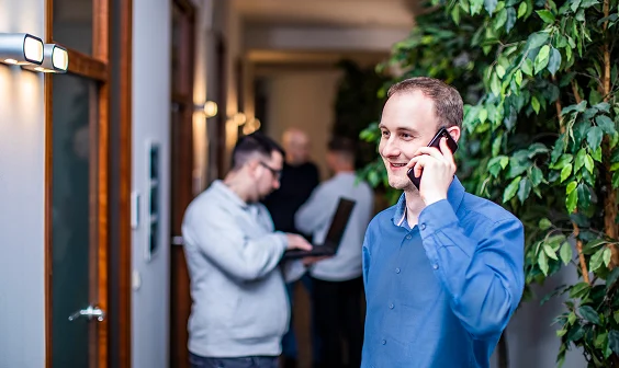 Toni in blauem Hemd telefoniert lächelnd im Büroflur, im Hintergrund Kollegen bei der Arbeit